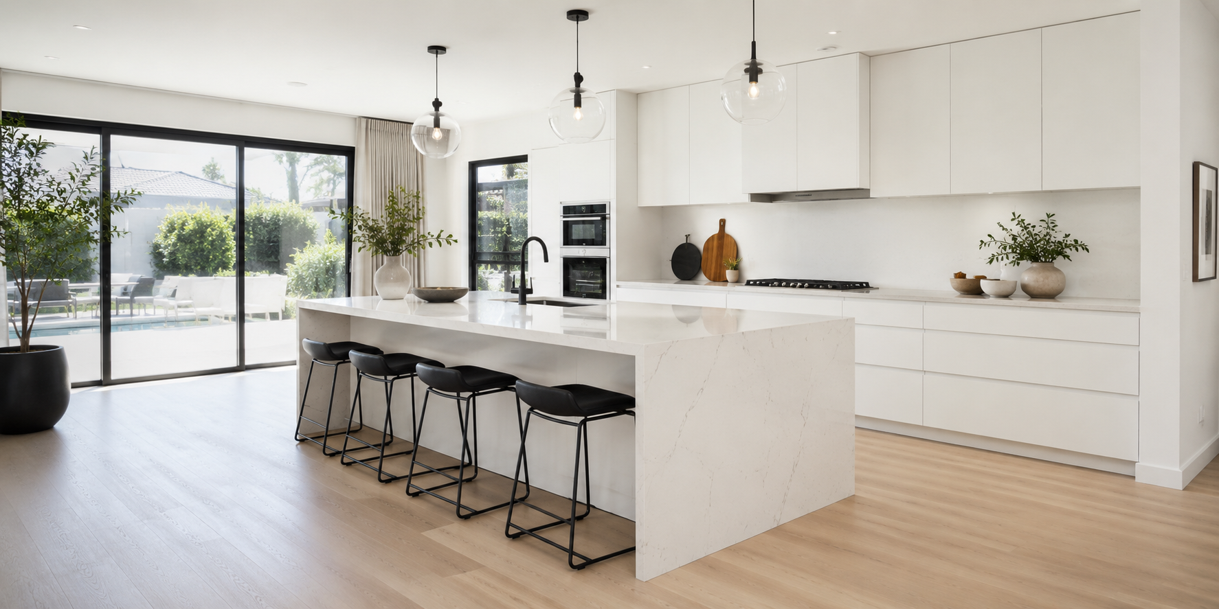 A minimalist kitchen with a white waterfall-edge island, integrated sink, matte black faucet, hidden cabinets, and bright natural light